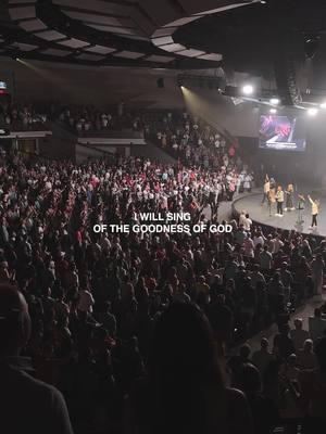 A wide shot captures a large auditorium filled with a crowd of people. The focus is on the stage where a group of people are standing. The lighting is dim with spotlights illuminating the stage. Text overlayed on the image read: 'I WILL SING OF THE GOODNESS OF GOD'.
