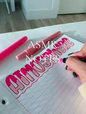 A close-up shot of a hand writing the word "AMAZING" in pink marker on lined paper. Two pink highlighters and a pen rest on the table around the notebook.