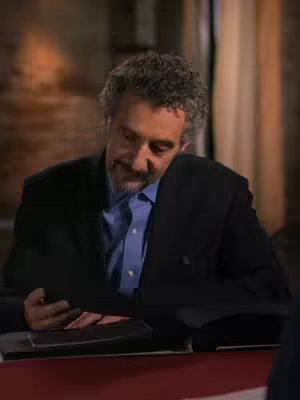 The image is a medium shot featuring a man with gray curly hair and a beard, wearing a blue button-down shirt and a dark blazer. He is seated behind a red surface, leaning forward as he looks down at something. He is holding a black object, possibly a document or book. The setting appears to be indoors, with a blurry background hinting at an office or a studio. The lighting is soft, and the overall composition suggests a moment of reflection or focused inspection. In the bottom part of the image is a blurry image of a person to the right.