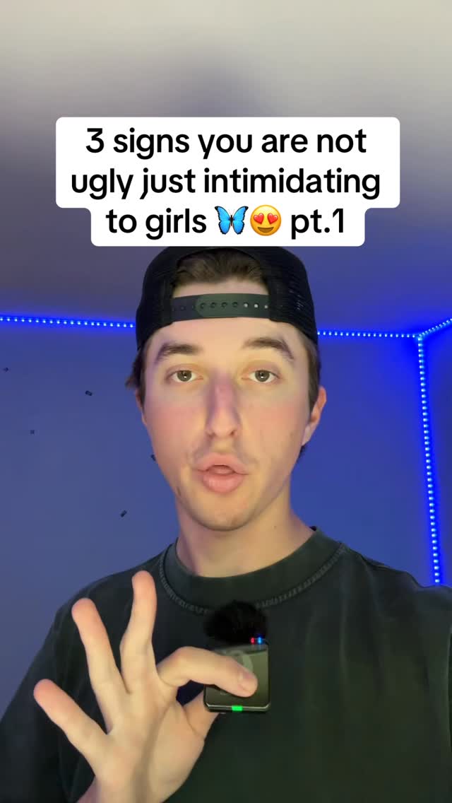 A selfie-style shot featuring a young man centered in the frame, with his face and upper body visible. He is wearing a black baseball cap and a dark gray t-shirt. A small microphone is clipped to his shirt. Above him, a text overlay proclaims: "3 signs you are not ugly just intimidating to girls." The background features a blue and white light pattern. The man is gesturing with his left hand, using his fingertips to create a gesture. His expression is neutral, and he has short brown hair