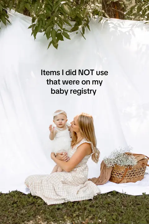 A vertical image featuring a woman seated, holding a baby. Both are dressed in light-colored, coordinated outfits. The woman is smiling, looking lovingly at the baby. The background is a simple white sheet, with a woven basket and a patch of green grass in the foreground. Text overlayed the image states, "Items I did NOT use that were on my baby registry".