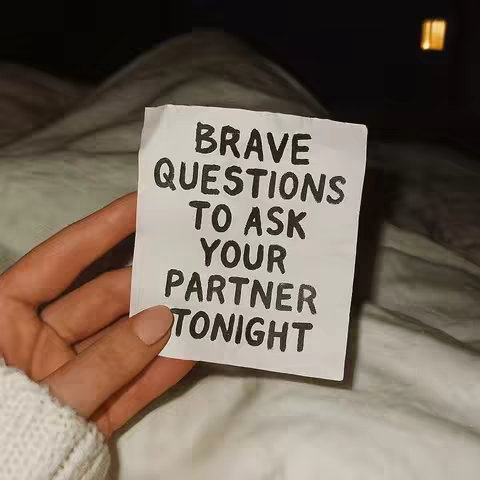 A hand holds a small white piece of paper with the text "BRAVE QUESTIONS TO ASK YOUR PARTNER TONIGHT" written on it in black, bold letters. The background is a blurred image of a dimly lit room, possibly a bedroom, with a bed visible. The lighting is soft and warm, with an overall intimate and cozy atmosphere.