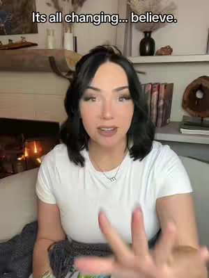 A woman is sitting on a couch, facing the camera, and speaking. The background includes a fireplace, shelves with decorative items, and books. The woman is wearing a white t-shirt and the hook text "Its all changing... believe." is written at the top of the image.