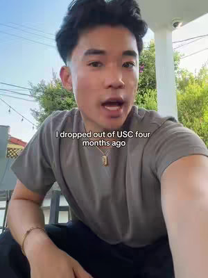 A young man is centered in a selfie video on a slightly overcast day. He is looking straight at the camera and is speaking, his mouth open as if he is making a statement. Text is overlaid on the image. The background shows a hint of foliage and the roof of what appears to be a patio or a porch with a ceiling fan. He is wearing a gray T-shirt and a gold necklace with a small square pendant. The lighting is diffused, creating soft shadows on his face. The overall composition is a standard selfie shot taken from a medium distance.