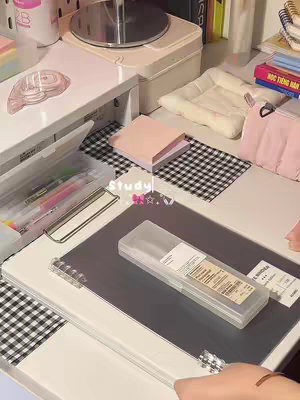 A top-down shot of a desk setup, featuring a black and white checkered pattern in the background and various stationery items. In the center is a clear case containing pens. Various other items are scattered around, including a box, note pad and more pens.