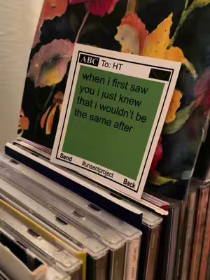 The image captures a collection of CDs in their cases, with a note card resting on top. The card is green with white borders and black text, it has a poetic message. In the background, out of focus, stand bright flowers.