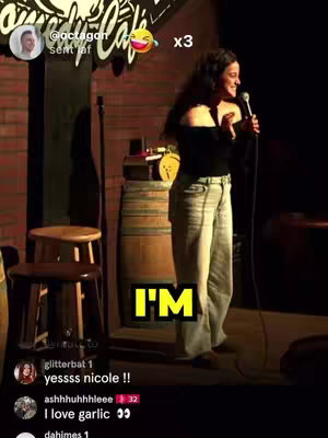 A woman stands on a stage with a microphone, looking slightly off-camera and smiling, with a yellow text box in the center with a word on it.