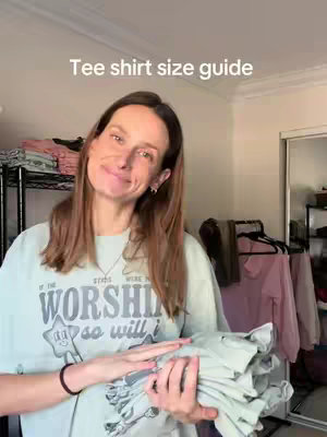 A woman is holding a stack of folded light green t-shirts. She is wearing a light green t-shirt and there is text overlayed in white on the shirt. The background has racks of folded clothing along with a closet in the background.
