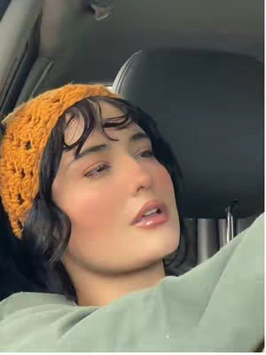 A woman with fair skin is seated in a car, seen from the chest up. She's wearing an orange, crocheted beanie and a green long-sleeved shirt. Her dark hair is styled with loose, wavy curls framing her face. The focus is on her face, specifically her expression, with hints of pink blush on her cheeks and full lips. The background shows the car's interior, including the driver's seat and headrest.