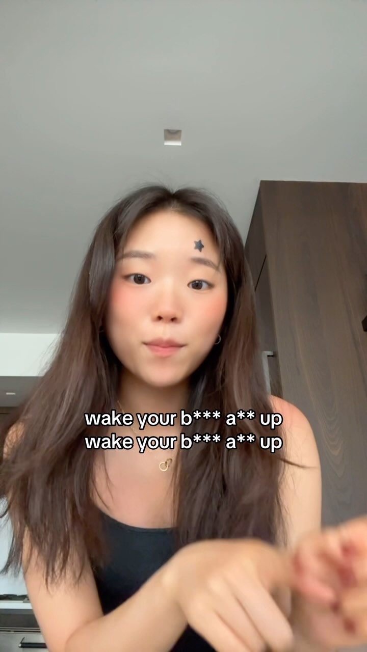 A young woman with long brown hair is the main focus. She wears a black top and a small star tattoo is visible on her forehead. Bold white text overlays the bottom portion of the image.