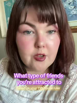 A selfie-style video shows a woman from the chest up with a medium shot. She has fair skin, bright pink blush on her cheeks, and dark red lipstick. Her dark brown hair has a short fringe, framing her face. Overlayed on her are the white text "What type of friends you're attracted to" in a purple gradient and under it "Based on your 11th house ruler" in white-yellow text. The background is out of focus with hints of artwork on the walls.