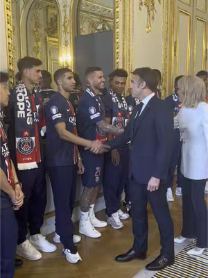 A group of men stand and shake hands in a well-lit room with ornate decorations. Several wear matching dark blue shirts and scarves, while others, including a man wearing a suit, shake their hands. The image is well-composed with a formal feel.