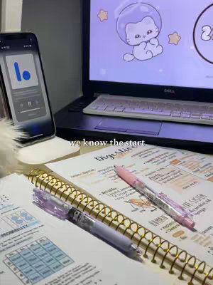 A well-lit workspace featuring a laptop with a cat-themed screensaver, an iPhone displaying a podcast app, study notes, and pens with a blurred background.