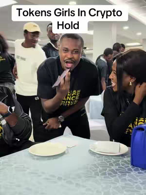 The image captures a scene involving a group of people indoors, seemingly at an event or meeting. A man in a black t-shirt with text is holding a card up near his mouth, his facial expression suggesting surprise or amusement. Beside him, a woman in a black top is smiling broadly. Additional people are visible in the background, which also includes a table with plates. The lighting seems consistent, with a soft, natural feel.