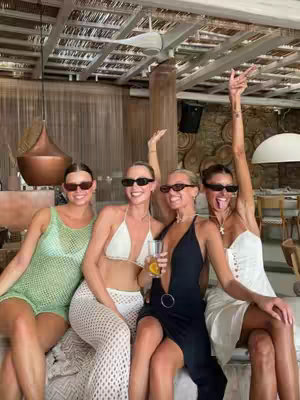 Four women are seated on a cushioned surface with a scenic background, likely a restaurant or outdoor lounge. All are wearing sunglasses, dresses/outfits suited for warm weather. One holds a cocktail and another is raising her arm in a pose. The overall mood is social and celebratory.