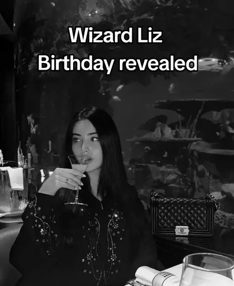 A black-and-white photo of a woman, presumably 'Wizard Liz', sitting in a restaurant or a similar upscale setting. She is holding a martini glass up to her lips, seemingly mid-sip. The background includes indistinct shapes and shadows, hinting at a bar or lounge setting, along with a part of a decorative aquarium. The composition is balanced, with the woman as the central focus.