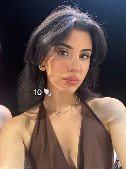 A young woman is centered in a selfie-style shot against a blurred, dark background. She has dark hair styled with layered bangs and is wearing a brown halter top and a delicate necklace. The text overlay reads "10" with a seashell emoji.