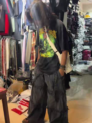 A person, presumably male, wearing a graphic print black t-shirt, heavily distressed dark gray pants, and dark shoes, is captured in a full-body shot. A white satchel bag is slung across the body. The background shows shelves of clothes and various items, giving the impression of what might be a shop interior. The lighting is natural, and the angle suggests an informal shot.
