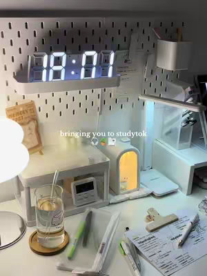 A well-lit, organized desk setting with an emphasis on study materials. A white pegboard is in the background, with an illuminated digital clock displaying '18:01'. Below the clock, a white shelf holds a small lamp and a white box of some kind. On the desk, there is a glass of water, pens, highlighters, notes, and a clear plastic pouch.