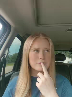 A young woman with long blonde hair is seated in the driver's seat of a car. She is wearing a blue t-shirt and has a thoughtful expression, with her index finger touching her lower lip, as if contemplating something. The car features a sunroof.