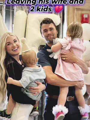A family portrait: a blond woman holds one child, smiling widely and a man with dark hair holds a second child, with a party hat, and giving him a kiss on the cheek, with a backdrop of white balloons.