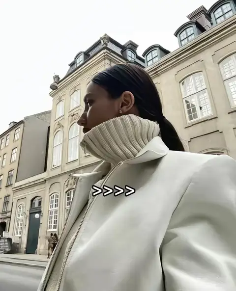 A woman in a white jacket and turtleneck looks to her left, set against a backdrop of European-style buildings. The buildings are ornate with many windows. The sky is overcast.