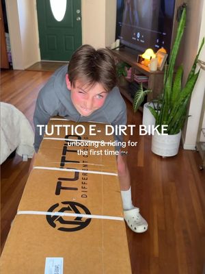 A young boy is holding a large cardboard box with the brand name 'Tuttio'. He is smiling and angled towards the camera over the box. Behind him is a blurred interior featuring a door and a plant.