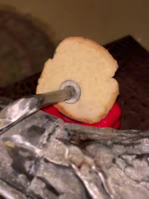 A close up shot showing a piece of bread pierced by a metal rod with a red object just behind the bread. The overall composition is dark with contrasting textures.