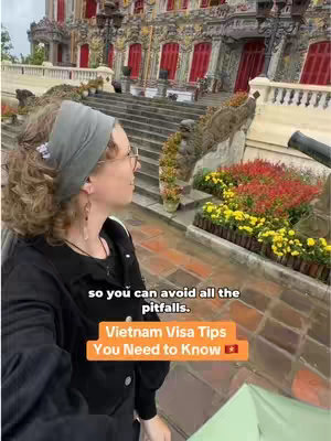 A woman stands facing away from the camera, looking towards a building in a foreign setting. The building has a detailed facade with red doors and windows. In the foreground is a flower bed with yellow and red flowers. The image has text overlays.