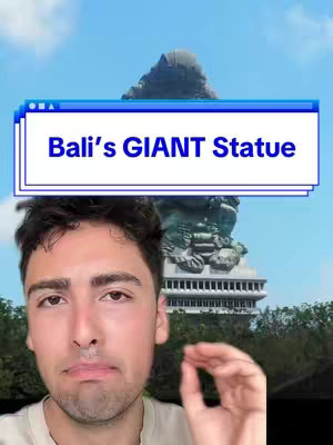The cover image features a young man in the foreground, partially obstructing the view of a large statue in the background. The man is fair-skinned with dark hair, and is looking at the camera. The backdrop is a clear blue sky with a large statue and a tall building. A text overlay in a blue rectangle reads, "Bali's GIANT Statue."