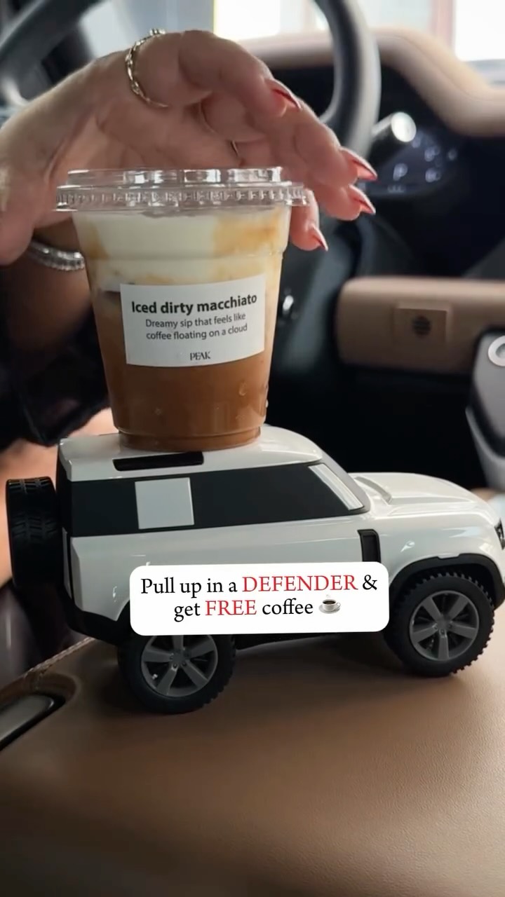 The image showcases a close-up of an iced coffee drink, with a label that reads "Iced dirty macchiato." The drink is placed on top of a small, white toy car resembling a Land Rover Defender. A hand with red-painted nails holds the drink, suggesting the setting is inside a car, likely the driver's seat. Below the car, a text layer on the image reads: "Pull up in a DEFENDER & get FREE coffee."