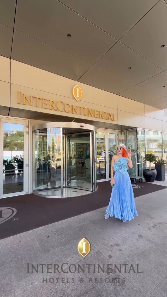 A woman in a blue gown stands in front of a revolving door entrance to the InterContinental Hotel. The InterContinental logo is visible, along with the hotel's name in gold lettering. The overall composition emphasizes luxury and elegance.