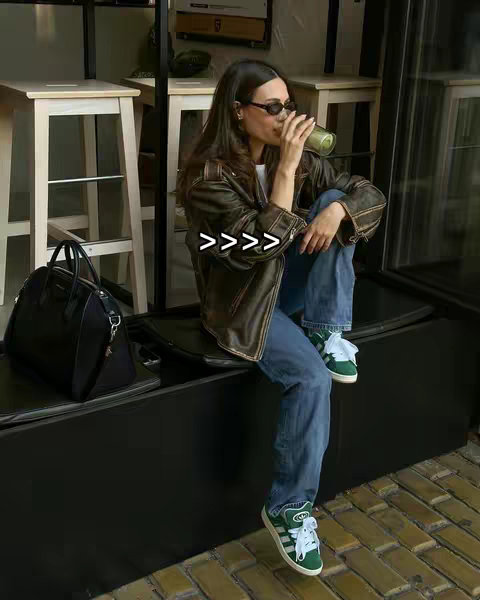 A woman in sunglasses, a brown leather jacket, and blue jeans sits on a black bench, holding a green drink. Behind her are white stools and a window.
