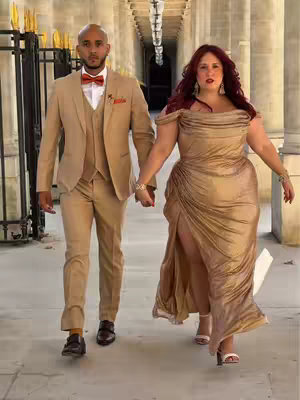 A man and a woman are walking hand-in-hand towards the camera. They are in a long hallway with arched ceilings and pillars. The man is wearing a tan suit and a bow tie. The woman is wearing a gold-colored gown. Both are looking at the camera.
