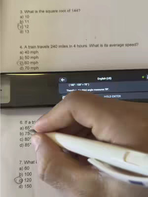 A hand holding a white pen is writing on a math test. The test paper is on a desk and a mobile phone is displaying a window, overlaid with some information and an English translation tag.