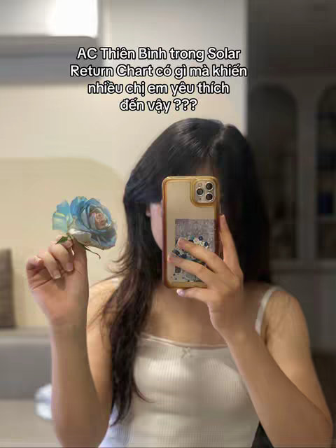 A woman holding a phone with a blue flower in front of a mirror. The background is a simple interior space, and the woman is wearing a white tank top.