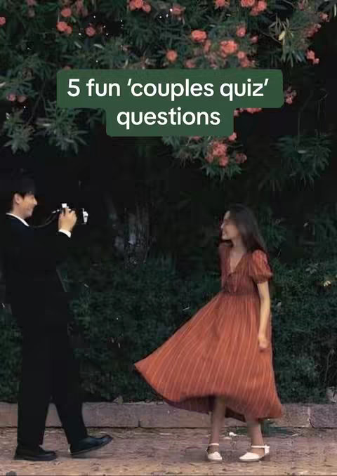 The image captures a couple in a natural outdoor setting. A man, dressed in a suit, is positioned on the left side, holding a camera. He appears to be taking a photo of a woman who is on the right side of the frame, wearing a flowy orange dress. The woman is in motion, possibly walking or dancing. Lush green foliage and blossoms form a backdrop, creating a soft, natural setting. Added text in the upper left corner displays the topic.