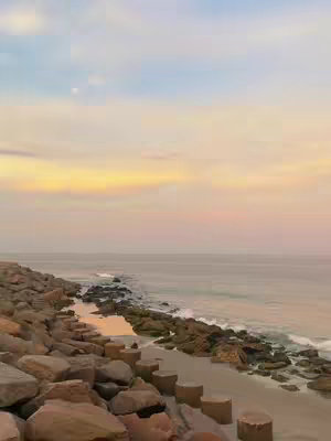A serene beach scene at sunset. The sky is a gradient of pale blue, yellow, and pink, with gentle clouds. The water is calm, reflecting the sky's colors. Rocks and sand line the shore, and there are some concrete blocks in the foreground. The overall feel is peaceful and picturesque.