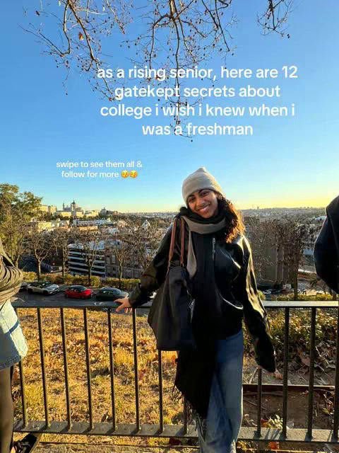 A young woman with long, dark hair and a beanie stands in front of a scenic outdoor background, looking towards the camera with a smile. She is wearing a black leather jacket, blue jeans, and a scarf. The background features a blurred cityscape and a bright blue sky.