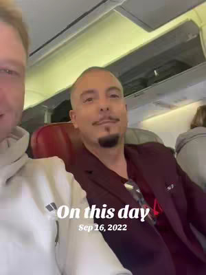 A selfie-style video shows two men inside an airplane during the day. The man on the right has a shaved head and a small goatee, wearing a maroon blazer over a black shirt. The other man is wearing a white hoodie. Overlay text indicates "On this day, Sep 16, 2022". The background suggests an airplane interior.