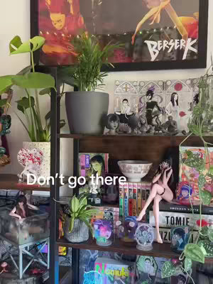 A well-lit, vertically oriented photograph of a bookshelf filled with anime and manga-related items. The shelves display various figurines, books, and framed art pieces, with plants strategically placed to add a decorative flair. The overall aesthetic is vibrant and reflects a collector's organized display.