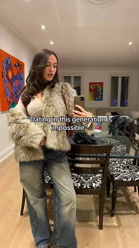 A young woman stands indoors, posing. She's wearing a furry, light-colored jacket, blue jeans, and a white top. She holds a phone, likely taking a selfie, with text overlayed on the image. The background includes a dining area with a glass table and ornate chairs, and artwork.