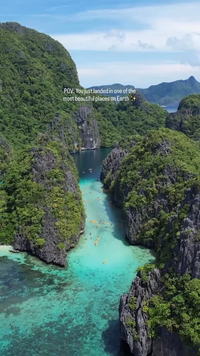 An eye-level shot captures a picturesque inlet, surrounded by towering, verdant cliffs. Crystal-clear, turquoise water fills the foreground, with numerous kayaks dotting the water. Above, a bright blue sky provides a backdrop, suggesting a sunny day. The composition highlights the natural beauty and clarity of the water.