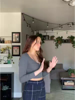 A woman stands in a well-lit room with white walls and a wood floor, gesturing with her hands. She wears a grey top and plaid pants. The background shows a sofa, framed pictures, and decorative string lights. A shelf with objects is on the left.