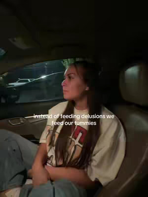 A woman is sitting in the front passenger seat of a car, looking to the side. She's wearing a light-colored t-shirt and jeans. Text overlayed on the image reads, 'instead of feeding delusions we feed our tummies.' The car interior is visible, with the seat and door frame in view. The background shows another car and a dark environment.