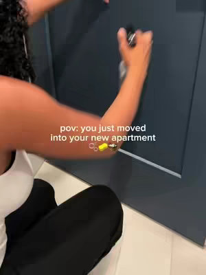 A woman is opening a door while holding keys. A text overlay reads 'pov: you just moved into your new apartment.' The background door is navy blue and the floor is light grey.