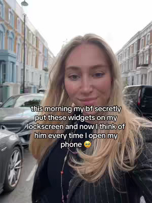 A selfie-style shot featuring a woman standing on a street with buildings in the background and vehicles parked nearby. The main focus is the woman, who is looking directly at the camera with a slight smile. Overlayed on the image is text describing a personal experience related to widgets on a phone, suggesting a tech-related or relationship-themed content.