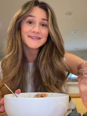A young woman with long, wavy brown and blonde hair, smiling and looking directly at the camera. She's holding a white bowl with food and a spoon, with a glass of pink liquid in the background. The lighting appears natural, and the setting suggests a domestic environment.
