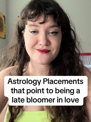 A young woman with long, wavy brown hair and red lipstick is the central focus. She smiles gently, with a white rectangular text overlay that has black text reading "Astrology Placements that point to being a late bloomer in love.". The background suggests a domestic setting.
