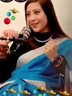 A woman with long, dark hair faces the viewer, smiling and speaking into a microphone. She wears a black top and a light blue and silver saree. Around her are several planet and star emojis in a playful design.